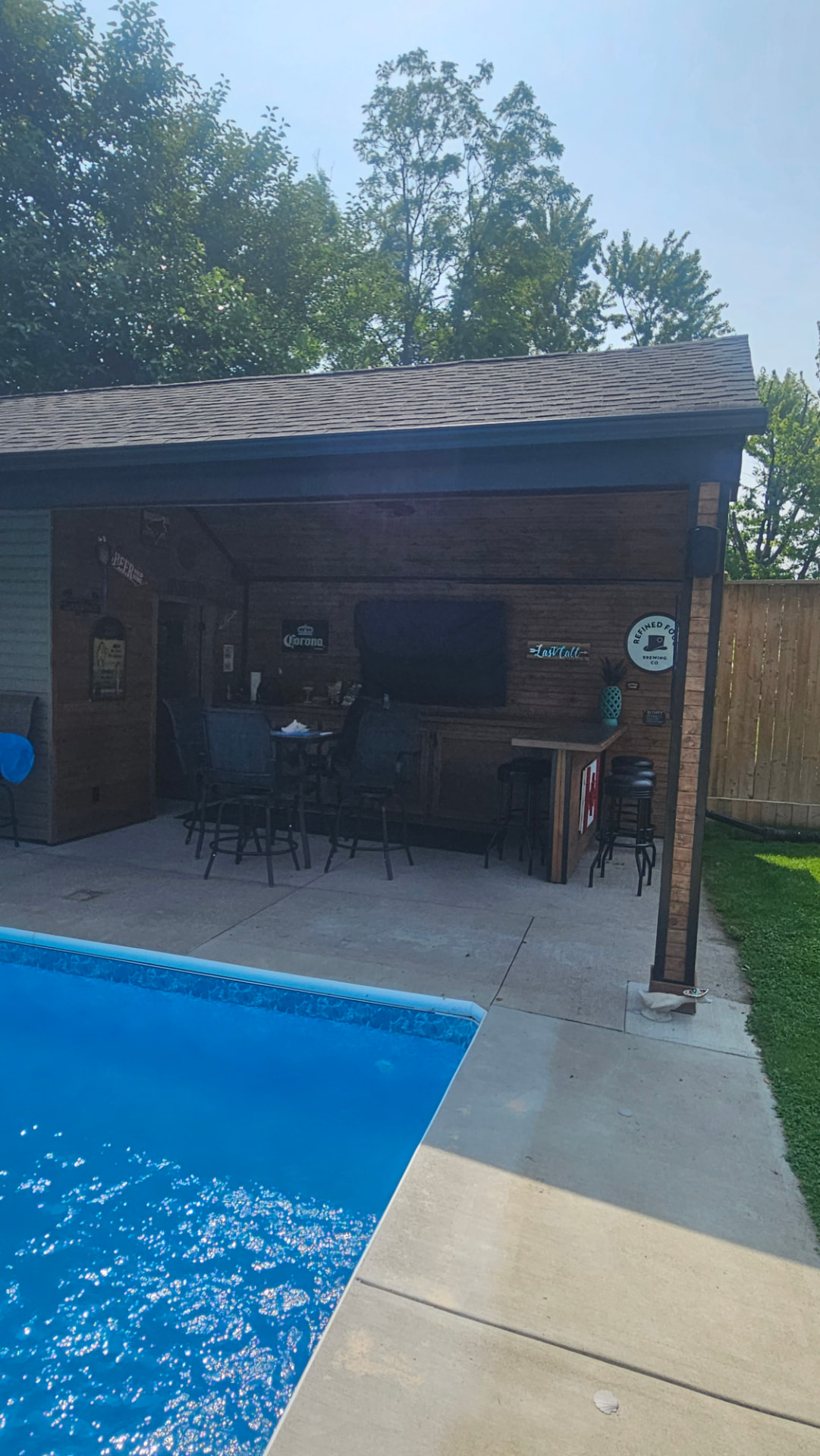 Poolside cabana with a covered outdoor bar, TV wall and bar seating beside an inground pool